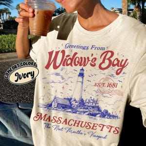 May include: An ivory t-shirt featuring a graphic print with the text Greetings From Widow's Bay, EST. 1681, and MASSACHUSETTS. The design includes a lighthouse, coastal buildings, birds, and a postmark stamp in red and blue ink colors.