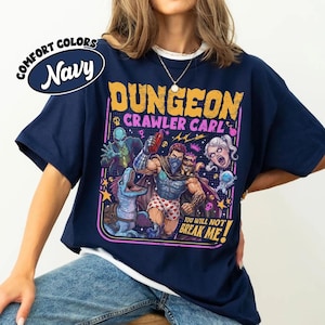 May include: A navy blue t-shirt featuring a graphic print with the text "Dungeon Crawler Carl" and "You Will Not Break Me!" The design displays a central muscular figure, a blue dinosaur, an alien, a cat, and a face, all in vibrant colours.