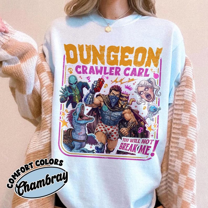 May include: A light blue sweatshirt featuring a graphic print with the text "DUNGEON CRAWLER CARL" and "YOU WILL NOT BREAK ME!". The design includes a central muscular figure, a blue alien, a dinosaur, and a floating head, all in vibrant colours.