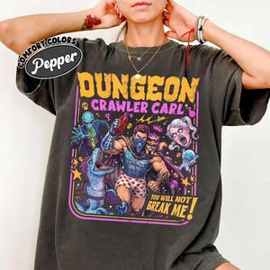 May include: A charcoal grey t-shirt featuring a graphic print with the text "Dungeon Crawler Carl" and "You Will Not Break Me!" The design includes a central muscular figure in heart-patterned boxers, a blue dinosaur, an alien, and a cat.