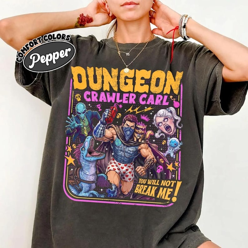 May include: A charcoal grey t-shirt featuring a graphic print with the text "Dungeon Crawler Carl" and "You Will Not Break Me!" The design includes a central muscular figure in heart-patterned boxers, a blue dinosaur, an alien, and a cat.