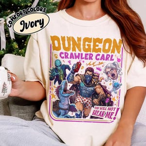 May include: An ivory t-shirt featuring a graphic print with the text DUNGEON CRAWLER CARL and YOU WILL NOT BREAK ME. The design includes a central muscular figure, a blue dinosaur, an alien, a cat, and a floating head, all in vibrant colours.