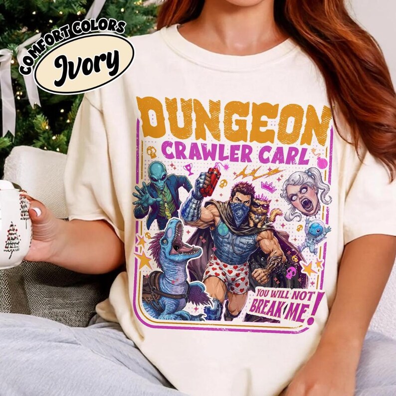 May include: An ivory t-shirt featuring a graphic print with the text DUNGEON CRAWLER CARL and YOU WILL NOT BREAK ME. The design includes a central muscular figure, a blue dinosaur, an alien, a cat, and a floating head, all in vibrant colours.