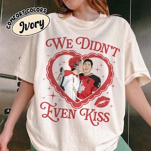 May include: Ivory-colored t-shirt with the text "We Didn't Even Kiss" in red script. The shirt features a heart-shaped graphic with two figures in hockey jerseys kissing. The shirt also has the words "Comfort Colors" in a circle.