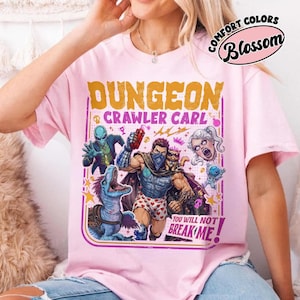 May include: A light pink t-shirt featuring a graphic print with the text DUNGEON CRAWLER CARL and YOU WILL NOT BREAK ME. The illustration depicts a central male figure in heart-patterned boxers, a blue dinosaur, an alien, a cat, and a floating head.