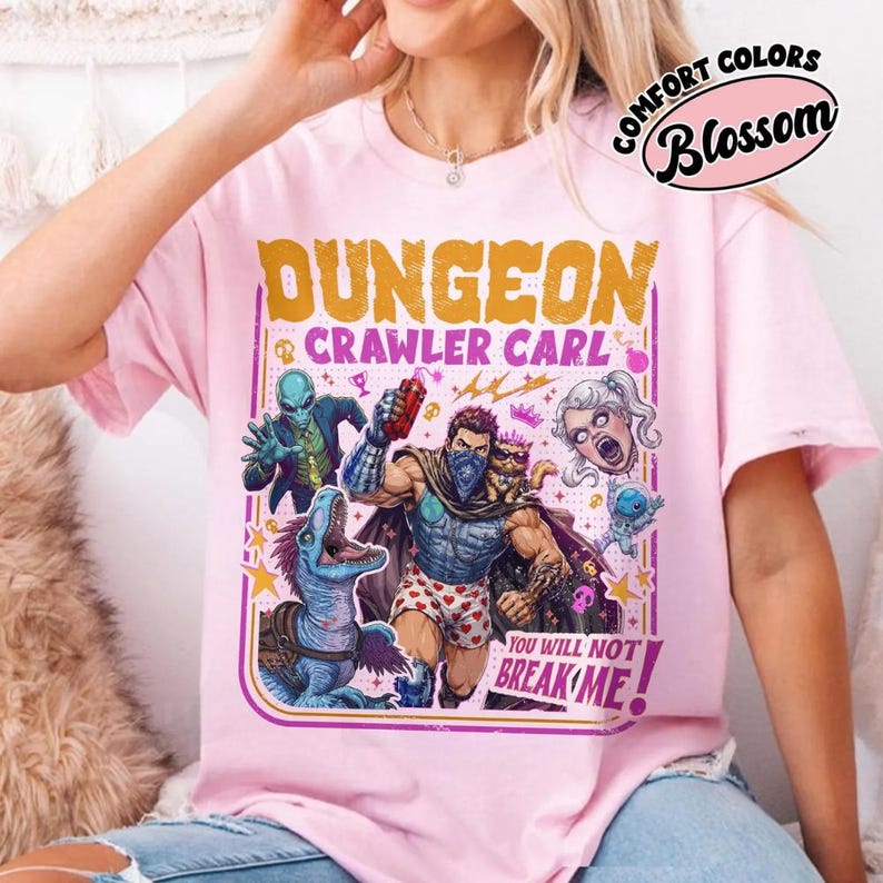 May include: A light pink t-shirt featuring a graphic print with the text DUNGEON CRAWLER CARL and YOU WILL NOT BREAK ME. The illustration depicts a central male figure in heart-patterned boxers, a blue dinosaur, an alien, a cat, and a floating head.
