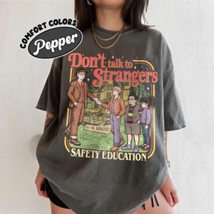 May include: Dark grey t-shirt featuring the text "Don't Talk to Strangers" and a vintage illustration of a man and children. The shirt also has the words "Safety Education" and "Comfort Colors Pepper".