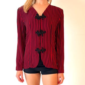 May include: Red and black striped long-sleeve blouse with black knot closures. The blouse is paired with black shorts and black ballet flats. The outfit is suitable for casual or semi-formal occasions.