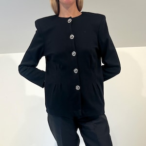 May include: A black suit consisting of a button-up jacket and matching pants. The jacket has silver-colored buttons and shoulder pads. The pants are straight-legged. The person is wearing black flats. The outfit is suitable for formal occasions.
