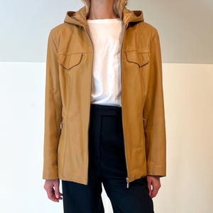 Puede incluir: Chaqueta de cuero color canela con capucha y dos bolsillos delanteros, usada sobre una camiseta blanca y combinada con pantalones negros de pernera ancha. La chaqueta tiene cierre de cremallera. El conjunto se completa con zapatos negros.