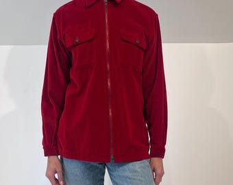 Puede incluir: Una camisa de pana roja con cremallera completa y dos bolsillos en el pecho. La camisa se combina con vaqueros de mezclilla azul claro. El modelo está de pie descalzo sobre un fondo blanco. El color de la camisa es un rojo intenso y rico.