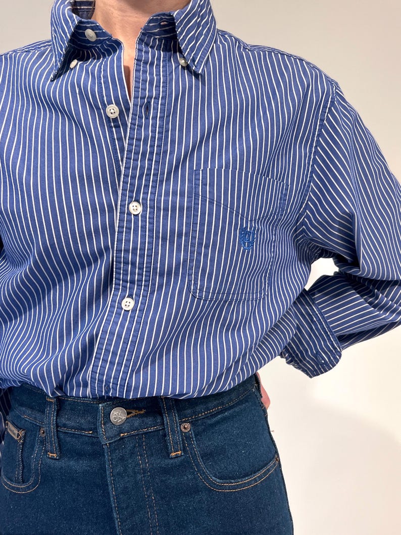Może przedstawiać: Koszula z długim rękawem zapinana na guziki, w niebiesko-białe pionowe paski, z kołnierzykiem typu button-down i pojedynczą kieszenią na piersi z wyhaftowanym niebieskim logo. Koszula jest wpuszczona w ciemnoniebieskie dżinsy z metalowym guzikiem.
