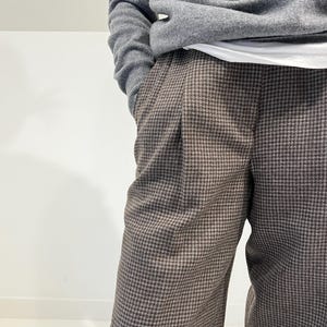 May include: A person wearing a gray sweater and brown houndstooth pants. The pants have a classic pattern and a pleated front. The sweater is a solid color and has ribbed cuffs.