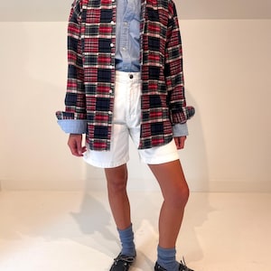 May include: A long-sleeve button-down shirt featuring a red, navy blue, and white plaid pattern worn over a light blue button-down shirt. The outfit includes white shorts, light blue ribbed socks, and dark navy blue leather boat shoes.