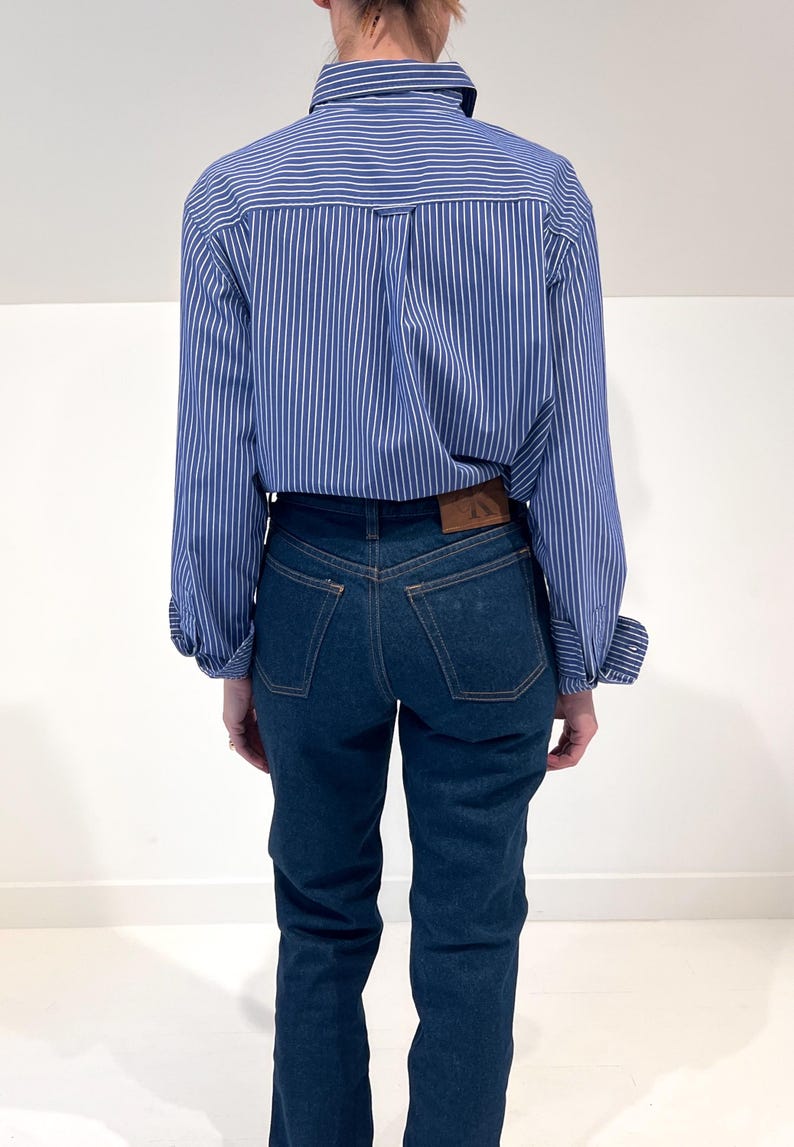 Może przedstawiać: Koszula z długim rękawem zapinana na guziki w niebiesko-białe pionowe paski, zestawiona z ciemnoniebieskimi dżinsami. Spodnie posiadają brązową sk&oacute;rzaną naszywkę z tyłu w pasie oraz dwie tylne kieszenie z widocznymi przeszyciami.