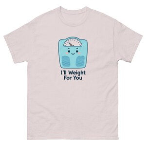 May include: Light pink t-shirt featuring a cartoon scale with a smiling face and the text "I'll Weight For You" in black. The scale is light blue with a white dial and two blue rectangles. The shirt is made of a soft material.