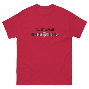 Puede incluir: Camiseta roja con la frase "IT'S JUST A PHASE" sobre una serie de ilustraciones de fases lunares en blanco y negro. La camiseta es de manga corta y cuello redondo.