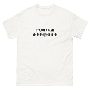Peut inclure: T-shirt blanc avec l'inscription "IT'S JUST A PHASE" au-dessus d'une série d'illustrations en niveaux de gris des phases de la lune. Le motif comprend la pleine lune et d'autres phases lunaires, créant un thème céleste.