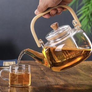 Könnte beinhalten: Eine Glasteekanne mit einem Holzgriff und einem eingebauten Sieb gießt Tee in eine Glas Tasse. Die Teekanne ist mit hellem braunem Tee gefüllt.