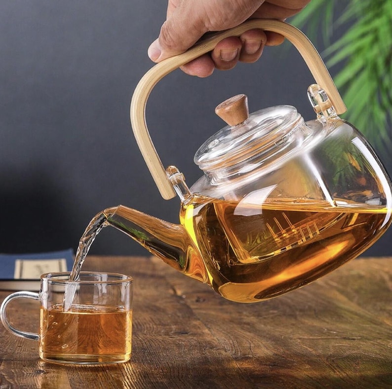 May include: A glass teapot with a wooden handle and a built-in strainer is pouring tea into a glass cup. The teapot is filled with a light brown tea.