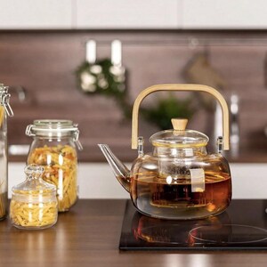 May include: A glass teapot with a wooden handle and a built-in strainer. The teapot is filled with brown tea and is sitting on a black electric stovetop.