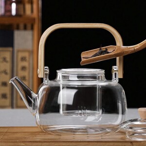 May include: A clear glass teapot with a built-in strainer and a wooden handle. The teapot is sitting on a wooden surface. A bamboo tea scoop is shown above the teapot.