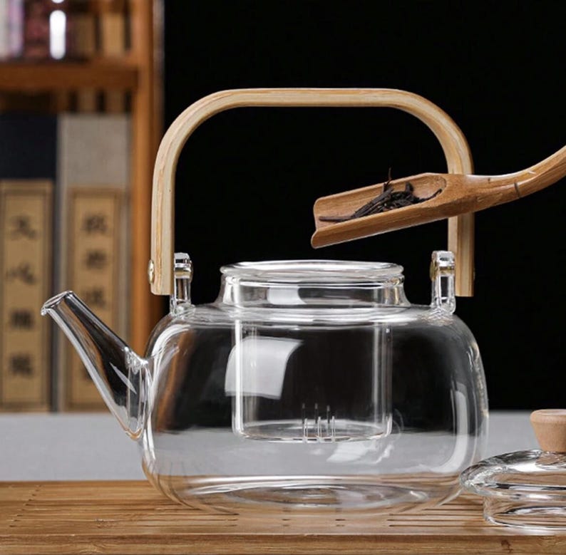 May include: A clear glass teapot with a built-in strainer and a wooden handle. The teapot is sitting on a wooden surface. A bamboo tea scoop is shown above the teapot.
