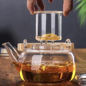 May include: A clear glass teapot with a wooden handle and a glass infuser. The infuser contains a yellow flower and is being lowered into the teapot. The teapot is filled with amber-colored tea.