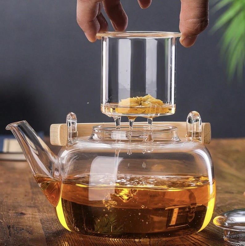 May include: A clear glass teapot with a wooden handle and a glass infuser. The infuser contains a yellow flower and is being lowered into the teapot. The teapot is filled with amber-colored tea.