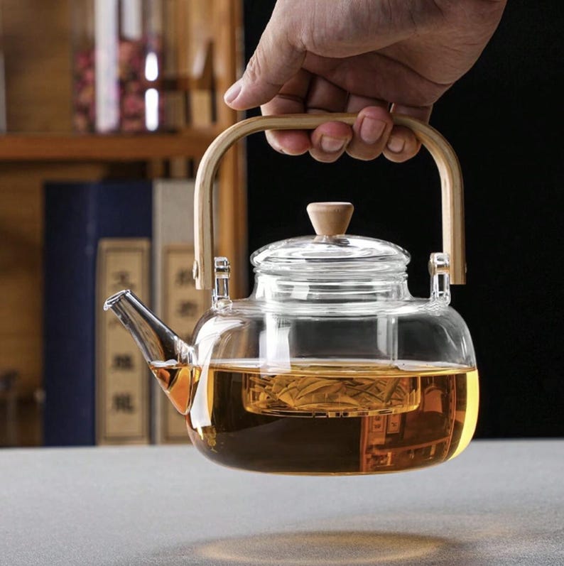 May include: A clear glass teapot with a wooden handle and a built-in strainer. The teapot is filled with a light brown tea.
