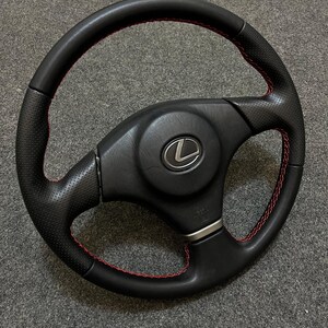 May include: Black leather steering wheel with red stitching and a silver Lexus logo.