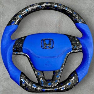 Puede incluir: Un volante de coche personalizado con un centro de cuero azul y empu&ntilde;aduras azules perforadas. El volante presenta un dise&ntilde;o de fibra de carbono con detalles azules y un emblema de Honda. El volante tiene controles integrados y el texto "SRS AIRBAG".