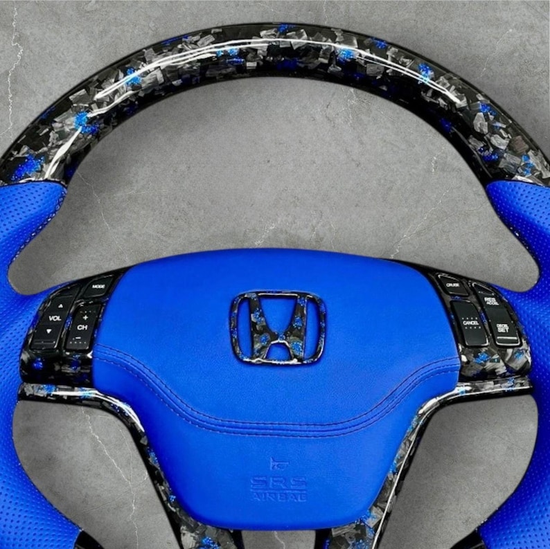 Puede incluir: Volante de coche personalizado con centro y detalles en cuero azul. La parte superior y los laterales presentan un dise&ntilde;o de fibra de carbono con motas azules. El volante tiene controles integrados y un emblema cromado de Honda en el centro.