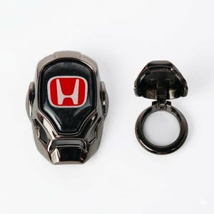 May include: A black and silver car key fob cover with a red and white Honda logo. The key fob cover is next to a matching key ring.