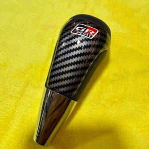 May include: A black and silver carbon fiber shift knob with a chrome base. The shift knob has the text "GR SPORT" in a red and white logo. The background is a bright yellow.