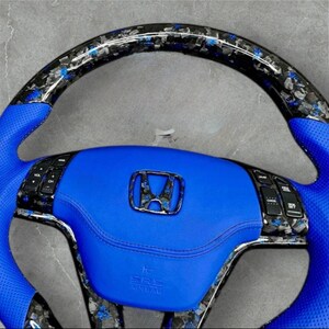Puede incluir: Volante de coche personalizado con una secci&oacute;n central y empu&ntilde;aduras de cuero azul brillante. La parte superior presenta un dise&ntilde;o de fibra de carbono con detalles azules. El emblema de Honda se muestra prominentemente en el centro.