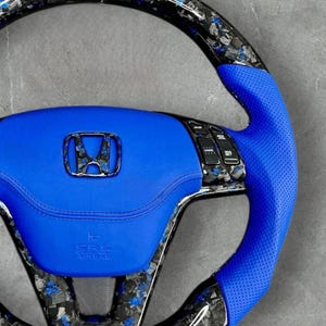 Puede incluir: Un volante de coche personalizado con un centro y agarre de cuero azul. El volante presenta un dise&ntilde;o de fibra de carbono con detalles azules y un emblema Honda cromado. El volante tiene botones de control integrados y las letras "SRS AIRBAG".