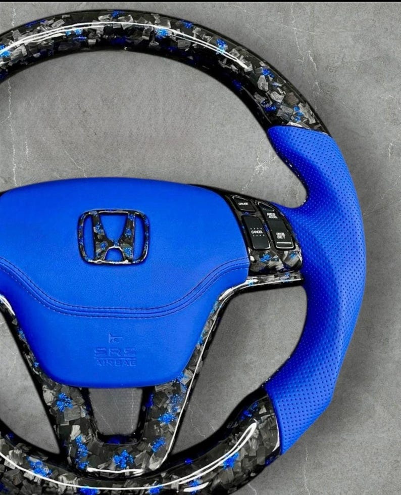 Puede incluir: Un volante de coche personalizado con un centro y agarre de cuero azul. El volante presenta un dise&ntilde;o de fibra de carbono con detalles azules y un emblema Honda cromado. El volante tiene botones de control integrados y las letras "SRS AIRBAG".