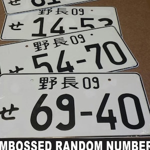 May include: A stack of white license plates with black embossed numbers and Japanese characters. The plates display different number combinations, including "14-52", "54-70", and "69-40". The text "EMBOSSED RANDOM NUMBERS" is at the bottom.
