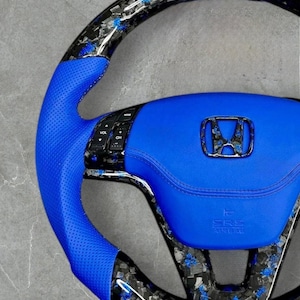 Puede incluir: Un volante de coche personalizado con un agarre de cuero azul y un borde de estilo fibra de carbono. El centro presenta una almohadilla de cuero azul con el logotipo de Honda y el texto SRS AIRBAG.
