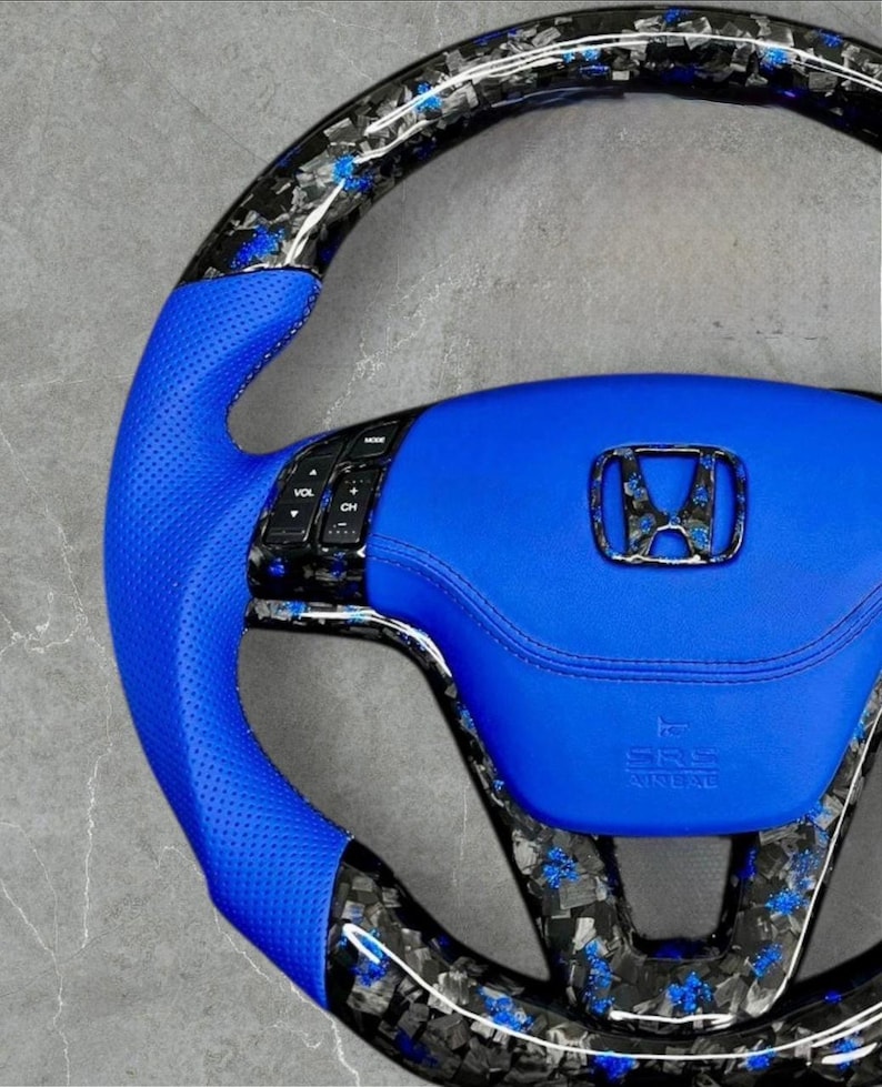 Puede incluir: Un volante de coche personalizado con un agarre de cuero azul y un borde de estilo fibra de carbono. El centro presenta una almohadilla de cuero azul con el logotipo de Honda y el texto SRS AIRBAG.