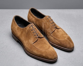 May include: A pair of tan suede leather oxford shoes featuring a rounded toe, a stitched apron front, and dark brown laces. The shoes have a low heel and a dark brown sole, with a visible logo embossed on the side of the right shoe.
