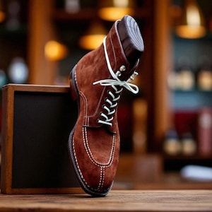 May include: A brown suede boot with white stitching and laces. The boot is displayed on a wooden surface with a wooden shoe form inside. The background is blurred, suggesting a store or workshop setting.