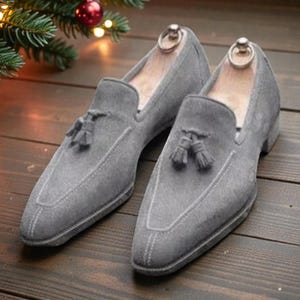 May include: A pair of gray suede loafers with tassel details. The shoes have a classic slip-on design and are displayed on a wooden surface. The image also includes a Christmas tree branch with red ornaments and lights.