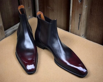 May include: A pair of polished leather Chelsea boots featuring a deep burgundy and black gradient finish. The boots have a sleek, pointed toe design, elastic side panels, and a low heel, showcasing a smooth, high-shine surface texture.