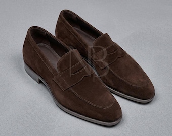 May include: A pair of dark brown suede penny loafers featuring a classic slip-on design with a horizontal strap across the vamp. The shoes have a rounded toe, a low heel, and a thin dark sole. A faint AB logo is visible on the side of the shoe.