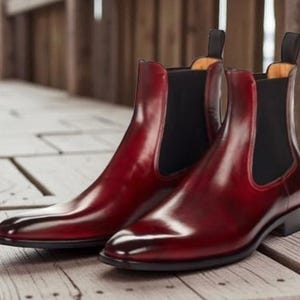 Op de afbeelding: Een paar bordeauxrode leren Chelsea boots met elastische zijpanelen en treklipjes. De laarzen hebben een glanzende afwerking en staan op een houten oppervlak.