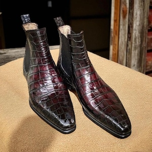 May include: A pair of burgundy Chelsea boots with a crocodile skin pattern. The boots have a glossy finish and black elastic side panels. The boots are displayed on a tan surface.