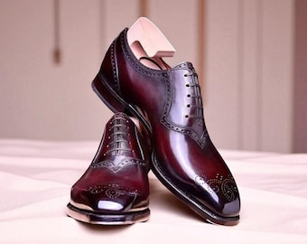 Puede incluir: Un par de zapatos de vestir de cuero burdeos con intrincados detalles brogue. Los zapatos se exhiben con una horma de madera dentro de uno de ellos. El cuero tiene un acabado brillante.