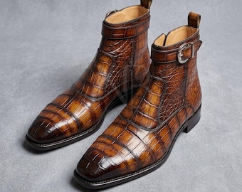 May include: A pair of ankle boots in a glossy brown finish with a textured crocodile skin pattern. Each boot features a side strap with a silver-toned metal buckle, a square toe, a pull tab at the heel, and a dark sole with a low block heel.